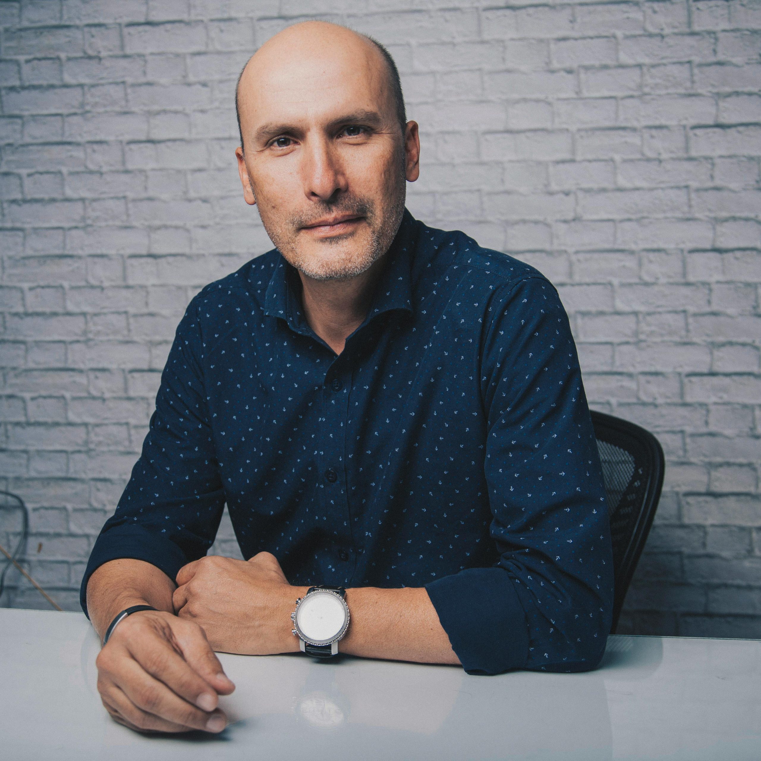 A confident middle-aged man sitting in a studio with a brick wall background.